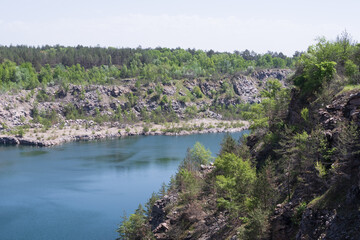 Fototapeta premium Flooded stone quarry with blue lake and rocky shoreline surrounded by green forest