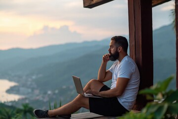 Obraz premium Man bearded using laptop on balcony with mountain view at sunset remote work