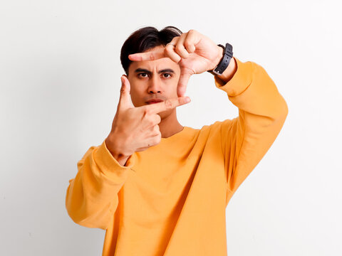 Focused Asian man making frame gesture with hands looking through fingers in yellow sweater isolated on white background.