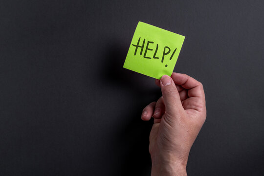 Male hand holding bright green sticky note asking for help on dark background