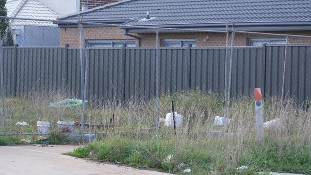 Unmaintained vacant block of land in a newly developed residential housing estate, overgrown with tall weeds in Melbourne Australia. The lot is fenced off with temporary fencing with scattered rubbish