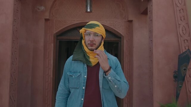 Young man wearing yellow green headscarf over face and glasses standing at carved terracotta riad arch