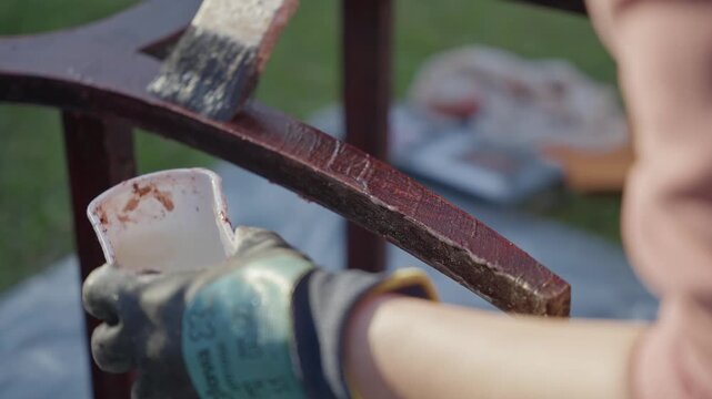 Person in gloves applying varnish removal chemical to weathered wooden chair frame outside