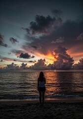 Fototapeta premium A Moment of Solitude at Dusk: A solitary figure stands on a sandy beach, gazing out at the tranquil ocean as the sky ignites with the fiery hues of sunset.