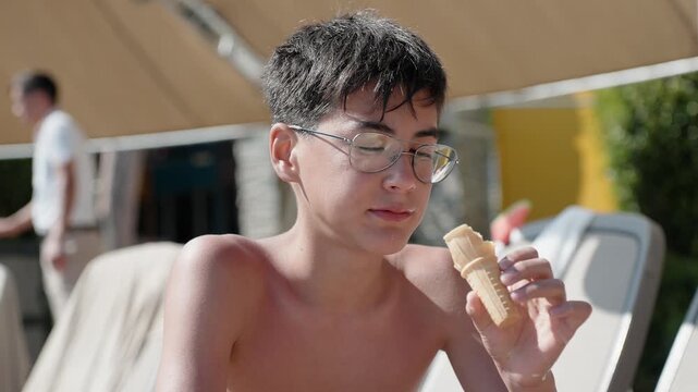 Male teenage holiday maker enjoying ice cream dessert while relaxing at sunny resort pool area. Boy eating dessert during summer holidays.