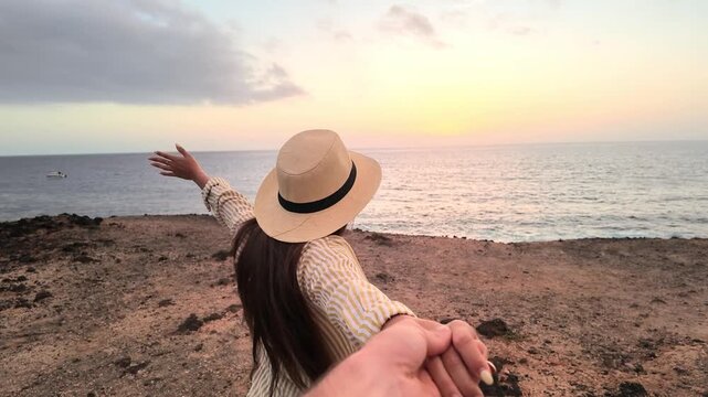 First-person perspective of a romantic young couple holding hands while walking toward the ocean, a beautiful woman leading her partner on a rocky beach to enjoy the amazing sunset view