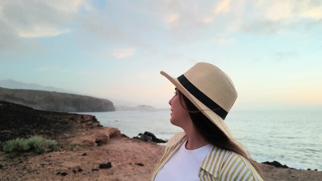 Beautiful young woman wearing a straw hat standing on a rocky cliff, smiling while enjoying the serene coastal view and the beautiful sunset over the ocean during her summer vacation