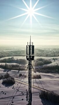 Telecommunications tower rising above snowy countryside under bright sun, drone vertical silhouette backlit