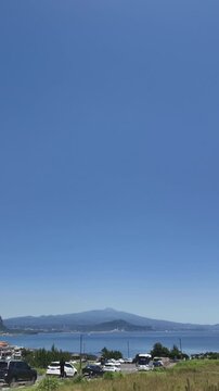 Clear blue sky and sea panorama of Jeju beach with distant mount Halla vertical stock video, Vertical shot of moving cars on coastal road with a scenic background of Sagye port and Hallasan
