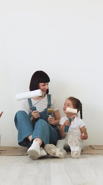 A mother and her young daughter sit on the floor, holding paint rollers and smiling at each other during a home renovation project