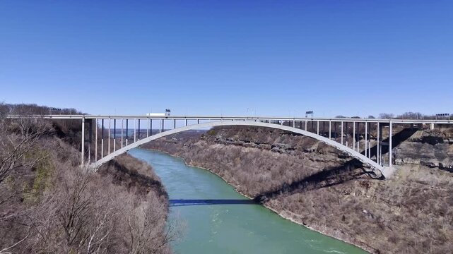 Aerial landscape featuring Lewiston Queenston Bridge crossing Niagara River gorge along Canada USA border beneath clear blue sky and rugged valley terrain.