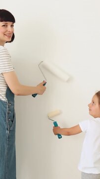 A mother and her young daughter happily paint a white wall together using rollers, with a ladder and painting pole nearby