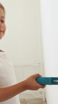 A young girl in a white t-shirt is painting a white wall with a roller, with a ladder in the background