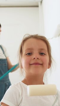 A young girl smiles at the camera while holding a paint roller, with her mother blurred in the background on a ladder also painting a wall