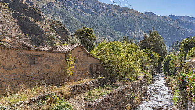 The mountain river flows through a narrow rocky riverbed. Along the shore there are old houses with masonry foundations and adobe walls. Tiled roofs. Green vegetation. Mountains, blue sky. Peru. Cusco