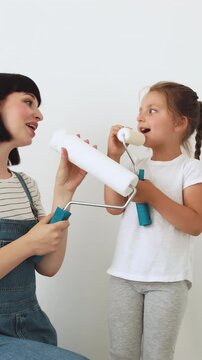 A mother and her young daughter are playfully holding paint rollers against a white wall, enjoying a moment of creative activity together