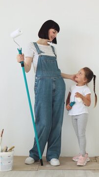 A mother and her young daughter stand side-by-side, holding paint rollers, ready to redecorate a room