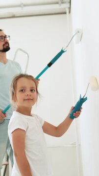 A father and his young daughter are painting a wall together with paint rollers in a bright room