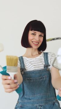 A smiling woman in denim overalls holds paint rollers and a brush, ready for home improvement projects