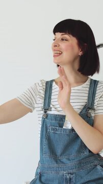 A woman in denim overalls is recording a video on her pink smartphone while standing near a ladder, possibly documenting a DIY project