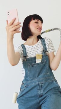 A woman in overalls sits on a ladder, holding a phone to record a video, with painting tools in her pocket