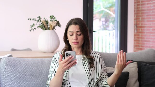 Distressed woman crying while having a difficult video conversation on her smartphone, sitting on the couch in her living room and gesturing with frustration and sadness, feeling heartbroken