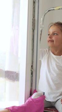 A mother wearing pink gloves cleans a window with a yellow cloth while her daughter sits on a ladder nearby