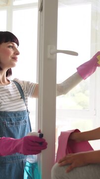 A mother and her young daughter are cleaning a window together, with the daughter on a ladder