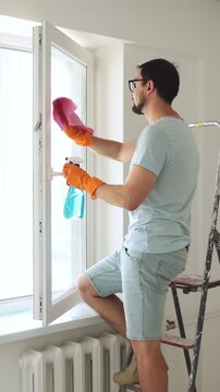 A man wearing orange gloves cleans a window with a spray bottle and cloth while standing on a ladder