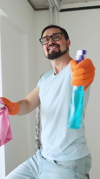 A smiling man wearing orange gloves holds a spray bottle and cloth, ready to clean a window in a bright room