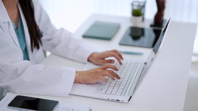 Asian women doctor medical professional typing laptop computer healthcare office desk clinic workspace telemedicine patient record documentation digital health