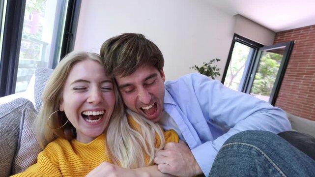 Joyful young couple laughing and having fun while taking a selfie video in their new apartment, running together and playfully falling onto the sofa in the bright living room