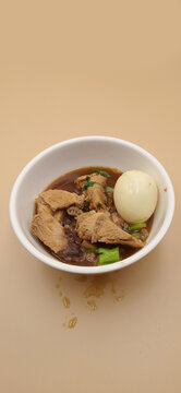 Braised pork leg with hard boiled egg in brown soup, Thai food known as Kha Moo Phalo, served in a white bowl on cream background.