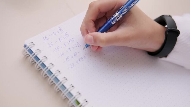 Hand writing math equations in dotted notebook with blue pen, solving arithmetic problems including negative numbers, educational activity, student homework, learning study session, school concept