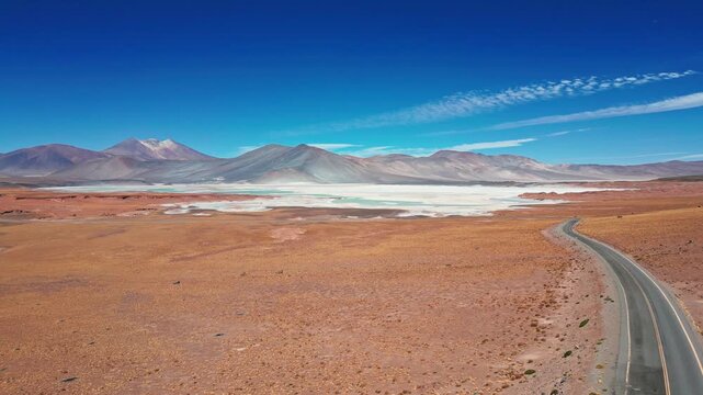 Dolly shot over winding paved road crossing red desert plain alongside iconic turquoise lagoon and white salt flat with multicolored volcanic peaks. Piedras Rojas, Chile