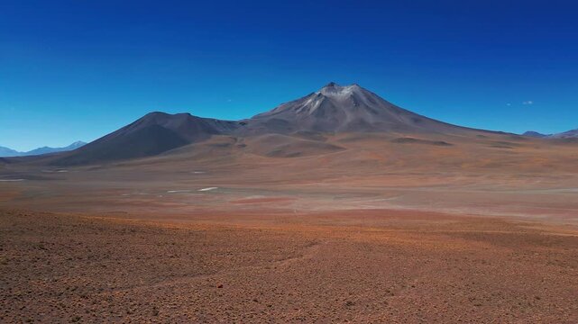 Low forward flight over vast red arid desert plain approaching a dramatic isolated volcanic cone rising sharply against deep blue cloudless sky in the altiplano