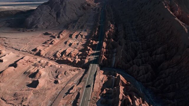 Tilt down aerial revealing car driving along straight highway cutting through dramatic eroded salt rock canyon between towering dark ridges. Valle de la Muerte, Chile
