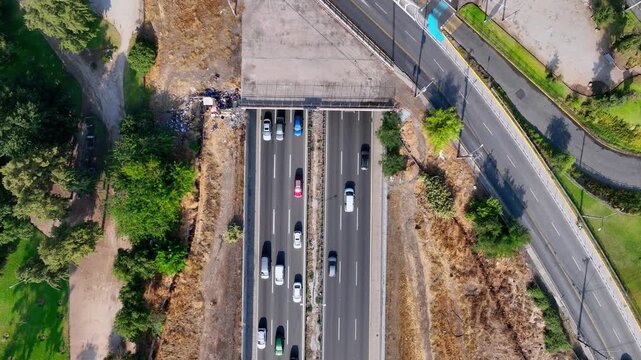 Top down ascending aerial shot over busy multi lane highway with cars flowing through urban underpass flanked by trees green verges and city streets on both sides