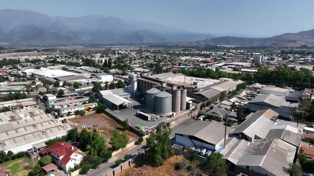 Orbit aerial shot over industrial grain silos and warehouse complex in a small urban town with residential neighborhoods and dramatic mountain range in the background under blue sky