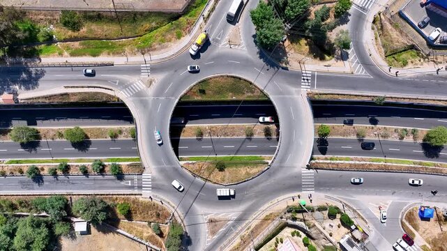 Aerial view over urban roundabout overpass with cars and trucks flowing through circular ring road above a highway underpass surrounded by trees and suburban streets