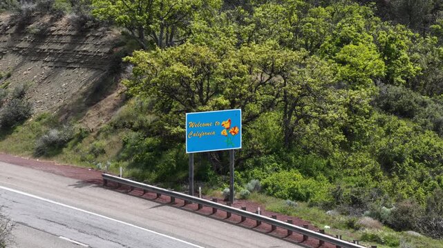 Welcome to California sign with poppy flowers close-up alongside highway guardrail, rocky layered slope and dense green oak trees under cloudy sky. California, USA. Aerial view.