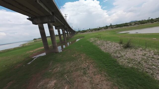 Thrilling low-altitude FPV drone shot flying under the concrete pillars of the Pa Sak Jolasid Dam railway bridge in Lopburi, Thailand. Highlights fast-paced maneuverability over green plains.