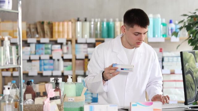 Young male pharmacist holds a box of pills and works at a computer in a pharmacy. Pharmacist offers customers high-quality medicines, tablets and ointments. High quality 4k footage