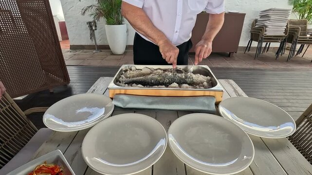 Timelapse footage of a waiter breaking and serving a whole salt baked fish at an outdoor restaurant table in Malaga Spain. The cook removes the salt crust and portions the fish onto white plates
