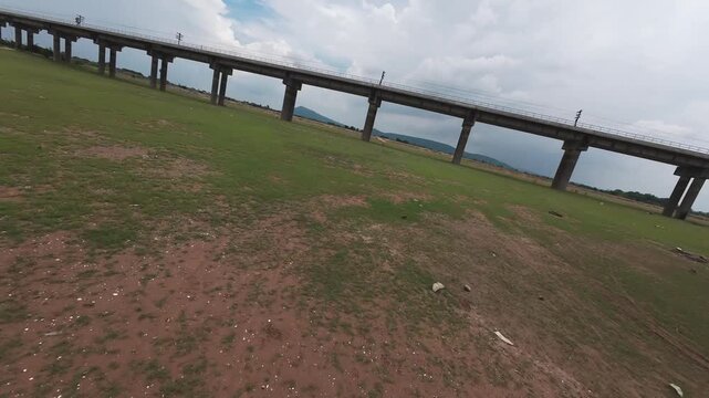 Thrilling low-altitude FPV drone shot flying under the concrete pillars of the Pa Sak Jolasid Dam railway bridge in Lopburi, Thailand. Highlights fast-paced maneuverability over green plains.