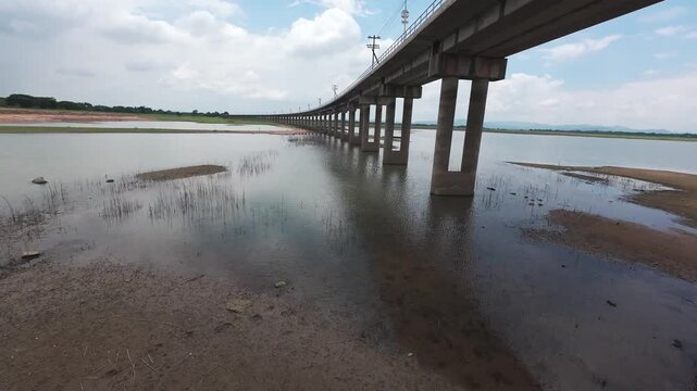 Thrilling low-altitude FPV drone shot flying under the concrete pillars of the Pa Sak Jolasid Dam railway bridge in Lopburi, Thailand. Highlights fast-paced maneuverability over green plains.