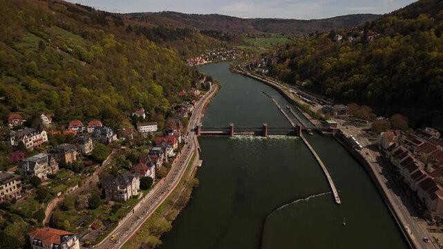 Aerial drone flight above the Neckar River in Heidelberg, Germany, showcasing a small dam, riverside roads, forested hills and scenic landscapes under bright sunshine in Baden-W&uuml;rttemberg, Europe.