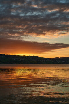 Golden sunset over calm lake with reflective water and dramatic cloudy sky above rolling hills