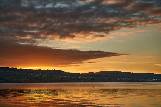 Golden sunset over a calm lake with village on rolling hills and dramatic cloud reflections