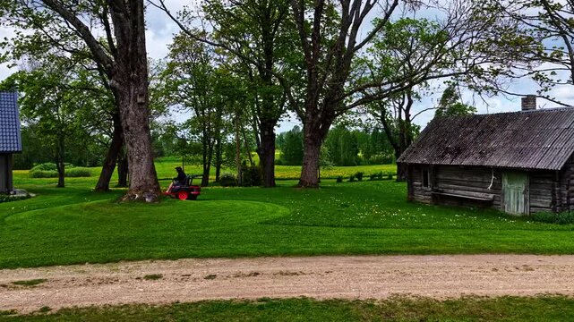 Person on red riding mower cutting lush green lawn between modern grey house and old rustic wooden barn with large trees in rural Jelgava Latvia on summer day
